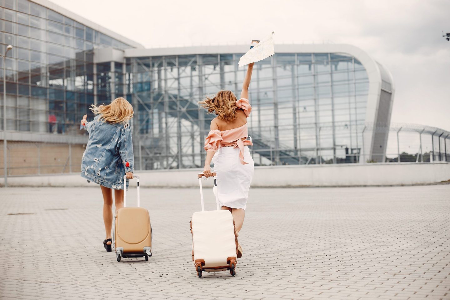 Two women running with suitcases toward a modern airport terminal for a last-minute trip.